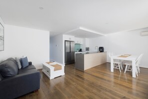 A modern kitchen with a central island, a dining area with white chairs, and a grey sofa with a throw pillow. - The Shoal at Shoal Bay (Shoal Bay)