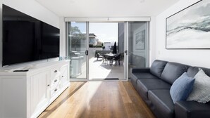Ein modernes Wohnzimmer mit einem Flachbildfernseher, einer Couch und einem Essbereich, der durch Glastüren sichtbar ist.