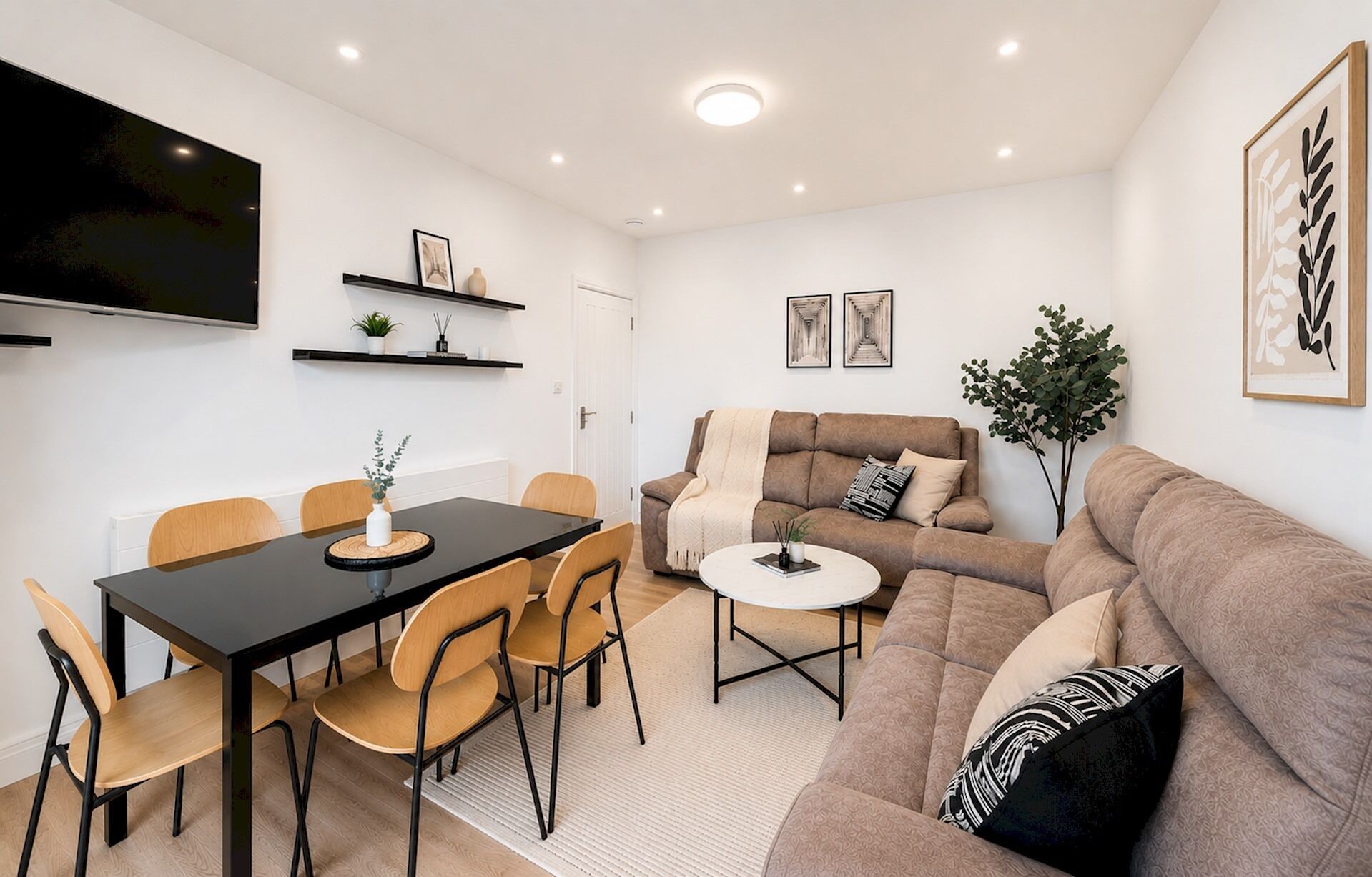 Un salon moderne avec un canapé, une table à manger et des chaises, un téléviseur fixé au mur et une petite plante.