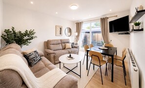 Ein modernes Wohnzimmer mit einer Couch, einem Esstisch mit Stühlen, einem an der Wand befestigten Fernseher und einem großen Fenster mit Vorhängen.