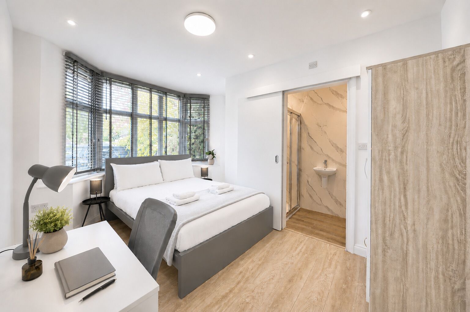 A modern bedroom with a bed, bedside tables, a desk with a lamp, a potted plant, and a shower area.