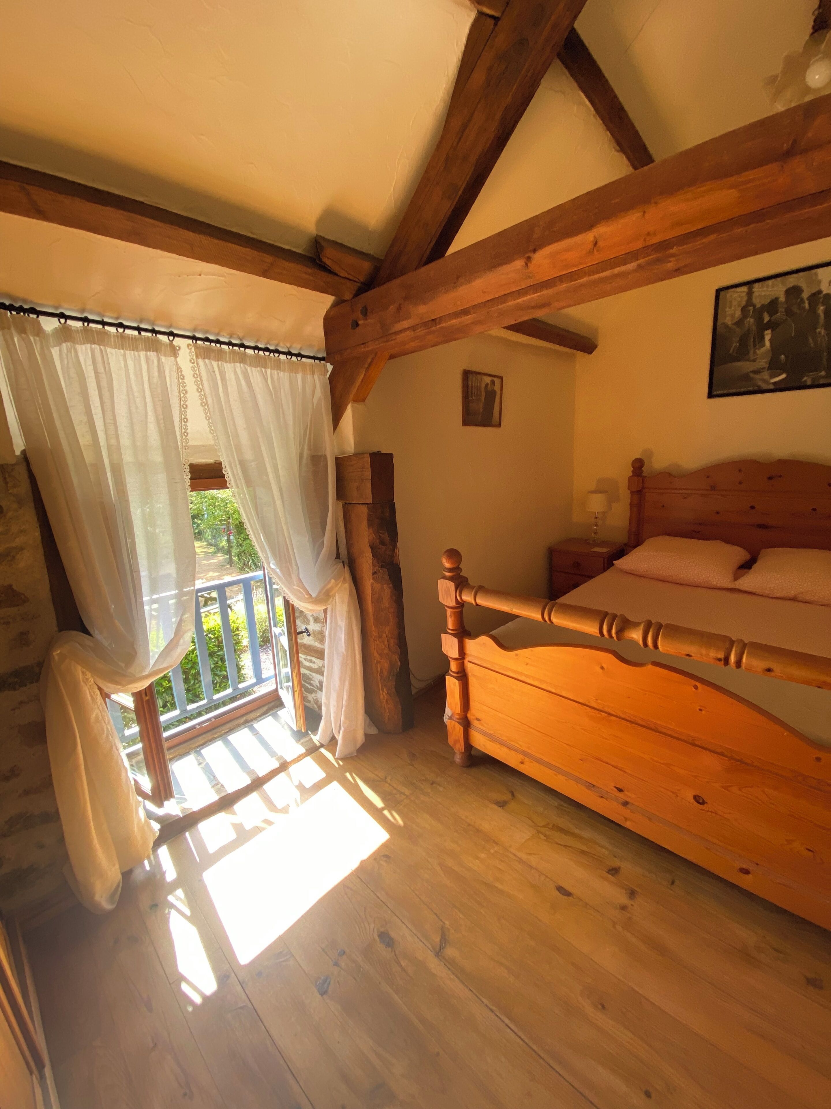 Een houten bed met hoofdeinde, een raam met dunne gordijnen en een ingelijst beeld aan de muur.