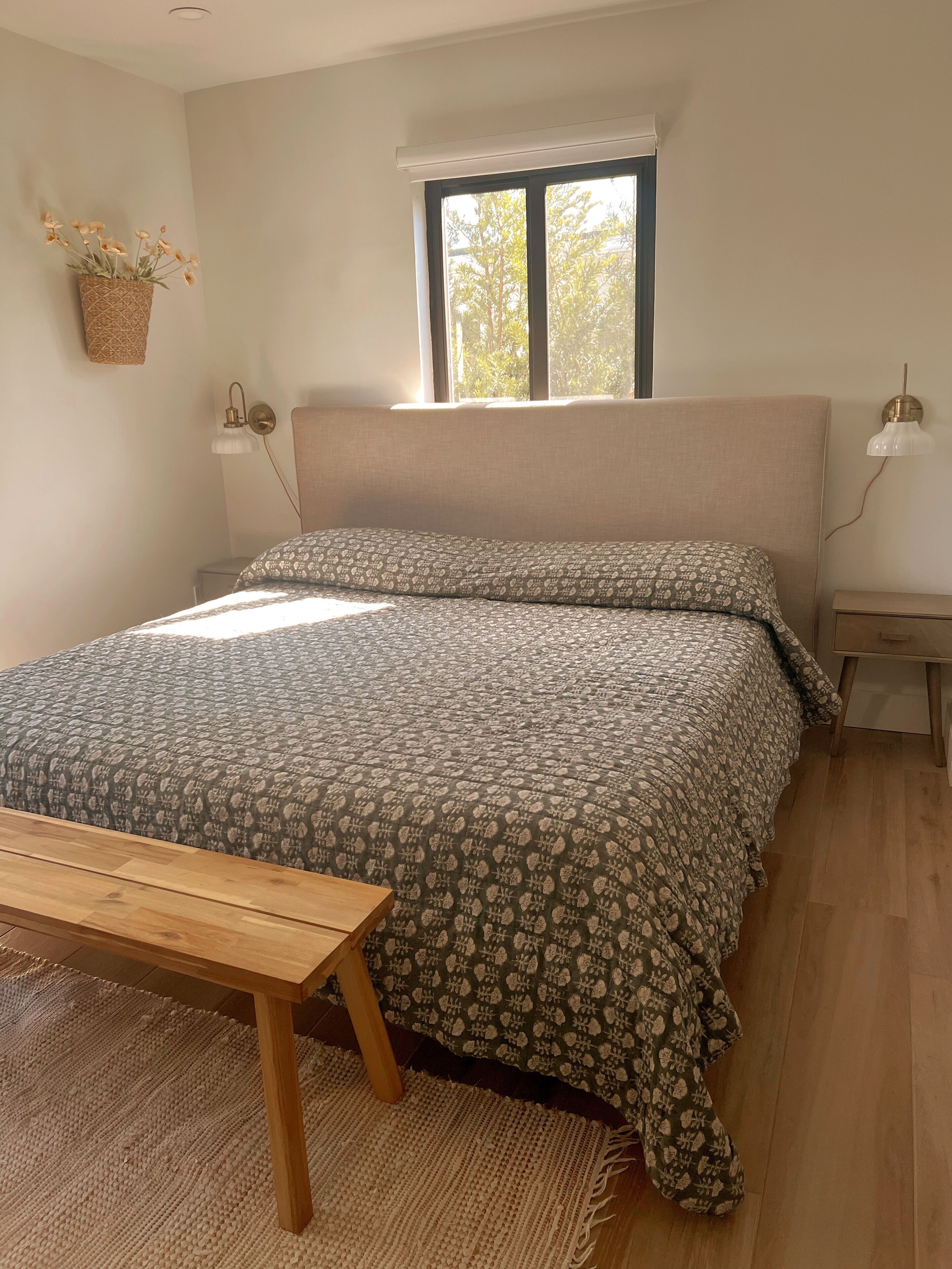 A bedroom with a bed, a bench, a window, and a wall-mounted lamp.