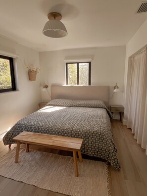 Ein Schlafzimmer mit Bett, Nachttischen, einem Fenster, einer hängenden Pflanze und einer Deckenleuchte.