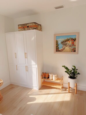 Ein Zimmer mit einem weißen Kleiderschrank, einem Schuhregal aus Holz, einem gerahmten Gemälde und einer Zimmerpflanze.