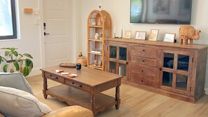 Uma sala de estar com uma mesa de centro de madeira, sofá de couro, televisão fixada na parede e um aparador de vime.