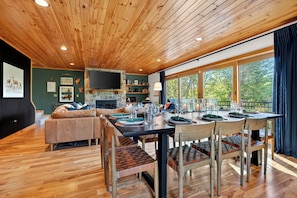 A wooden ceiling, a stone fireplace, a dining table with chairs, a sofa, and a TV mounted on the wall. - Ultimate Family Adventure Basecamp: Golf Sim, Rock Wall, Hot Tub, Sauna & More (Bethel)