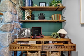 A room with a stone wall, wooden shelves with books and decorative items, a table with a lamp, and a suitcase.
