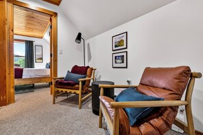 A cozy room with a bed, two wooden chairs, a small table, and a lamp. There are two framed pictures on the wall.