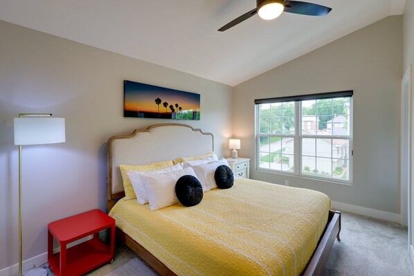 A bedroom with a bed, a nightstand, a red side table, a ceiling fan, a window with a view, and a painting on the wall.