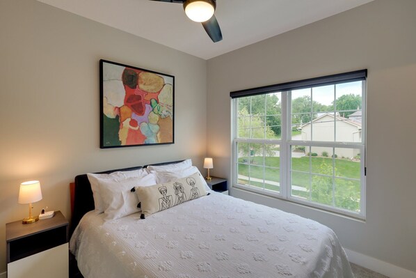 A bedroom with a bed, a window, a ceiling fan, and a painting on the wall.