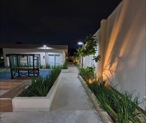 A narrow pathway flanked by greenery and a pool on one side, leading to a building with a door and windows.