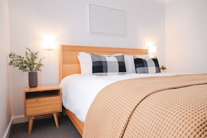 Ein ordentlich eingerichtetes Schlafzimmer mit einem hölzernen Kopfteil, einem Nachttisch mit Pflanze und einem großen Bett mit einer beigen Decke.