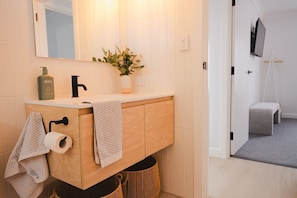 Ein Badezimmer mit einem Holzbadezimmermöbel, einem Spiegel, einem Waschbecken, einem Toilettenpapierhalter, einem Handtuch, einer Pflanze und einem an der Wand montierten Fernseher.