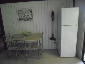 A small kitchen with a white refrigerator, a table with chairs, a bowl, and a framed picture on the wall.