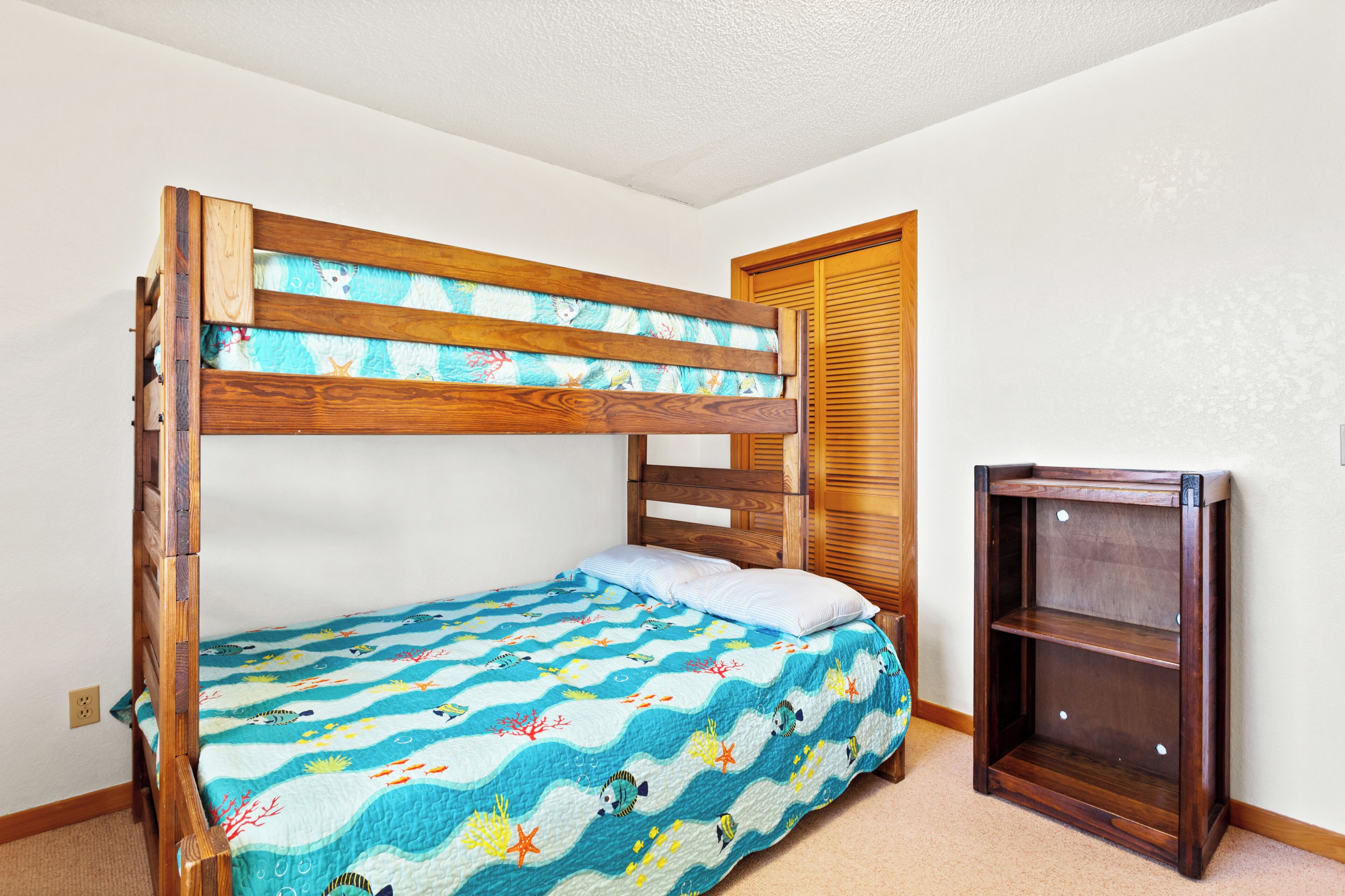 Una cama litera con un edredón de temática marina en azul, un cabecero de madera y una pequeña estantería de madera a su lado.