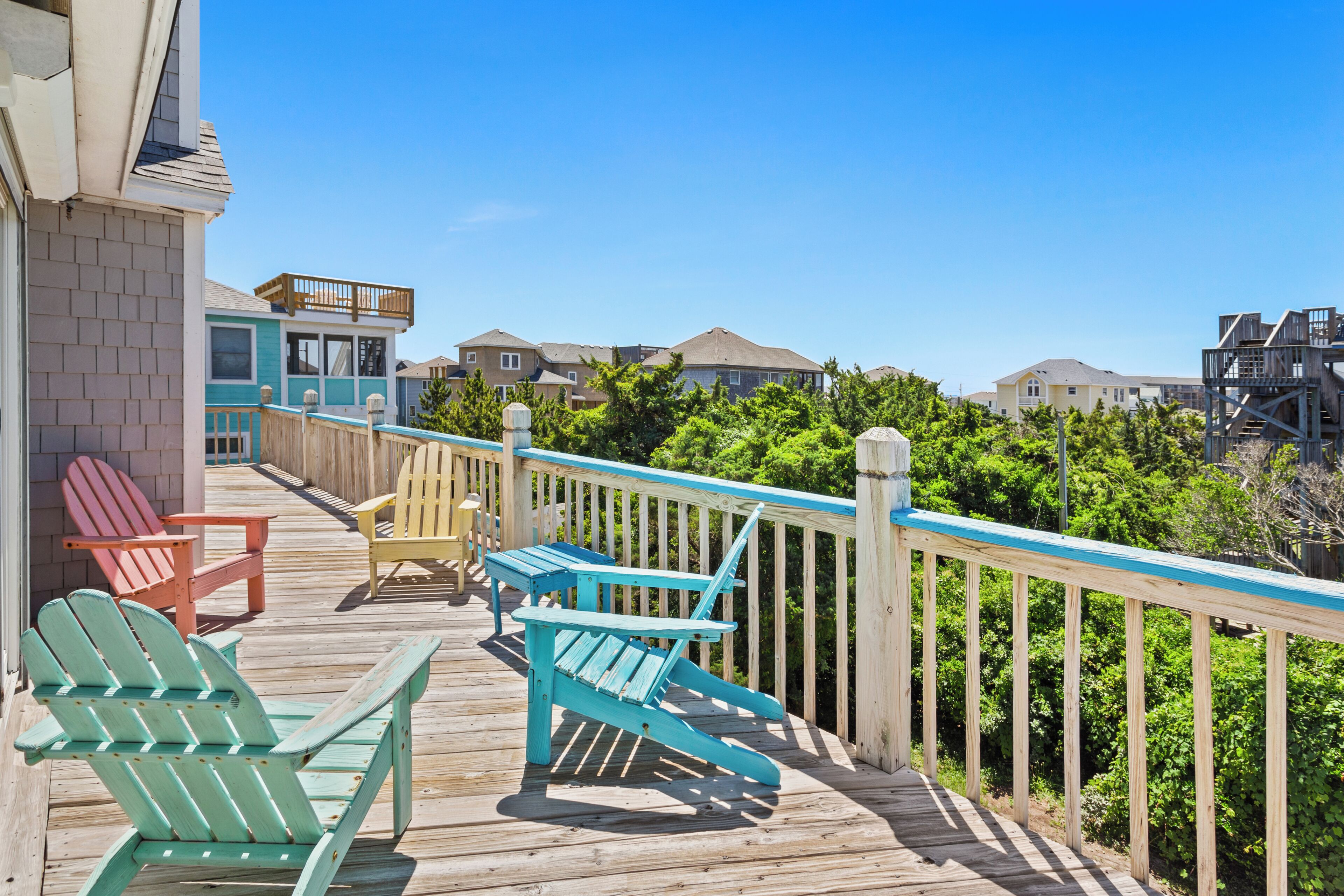Une terrasse en bois, agrémentée de chaises Adirondack, d’un banc et d’une rambarde, qui offre une vue sur un quartier résidentiel avec ses maisons et sa verdure.