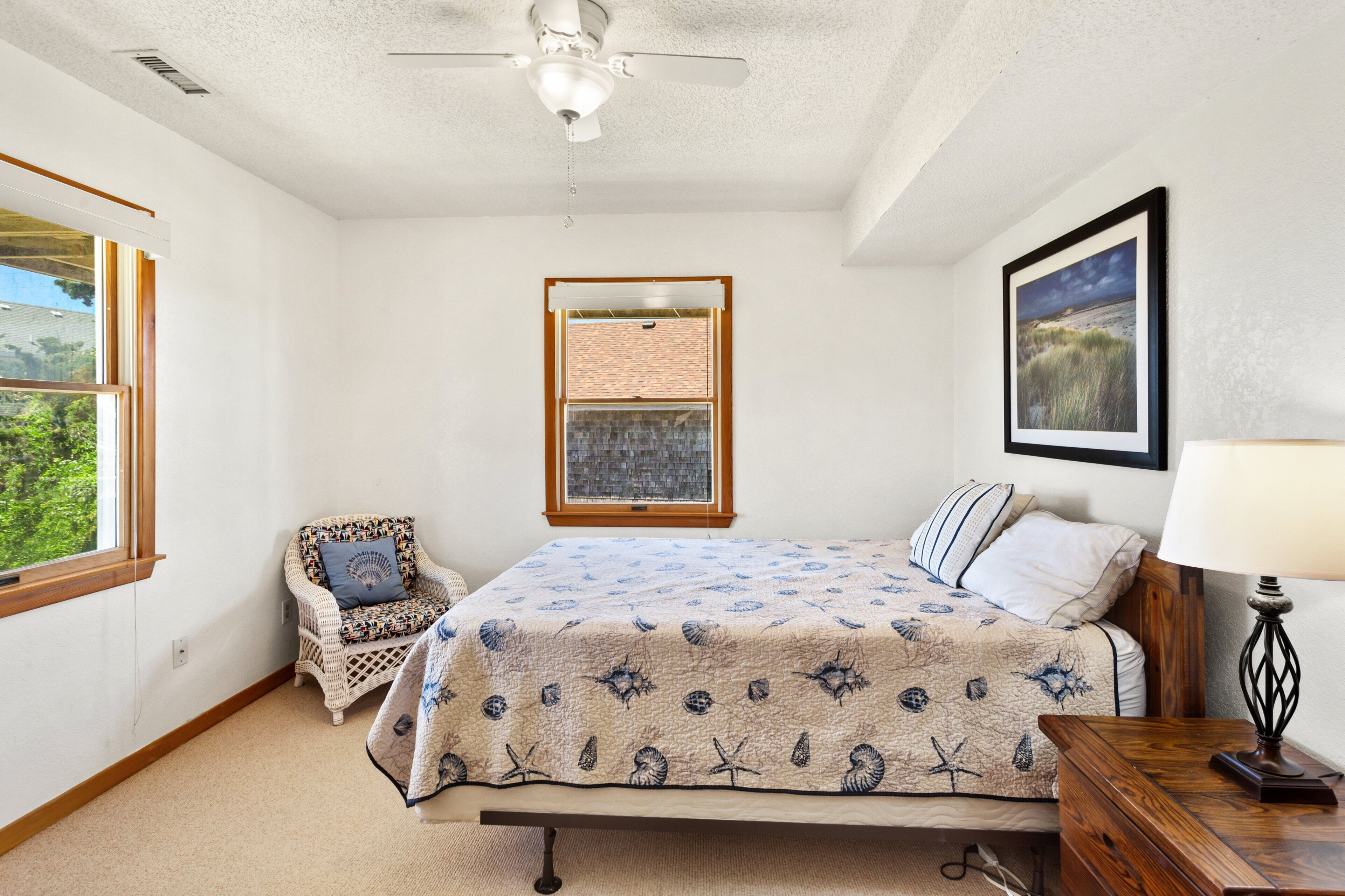 Une chambre à coucher comprenant un lit, une chaise, un ventilateur de plafond, une fenêtre, un tableau encadré et une lampe.