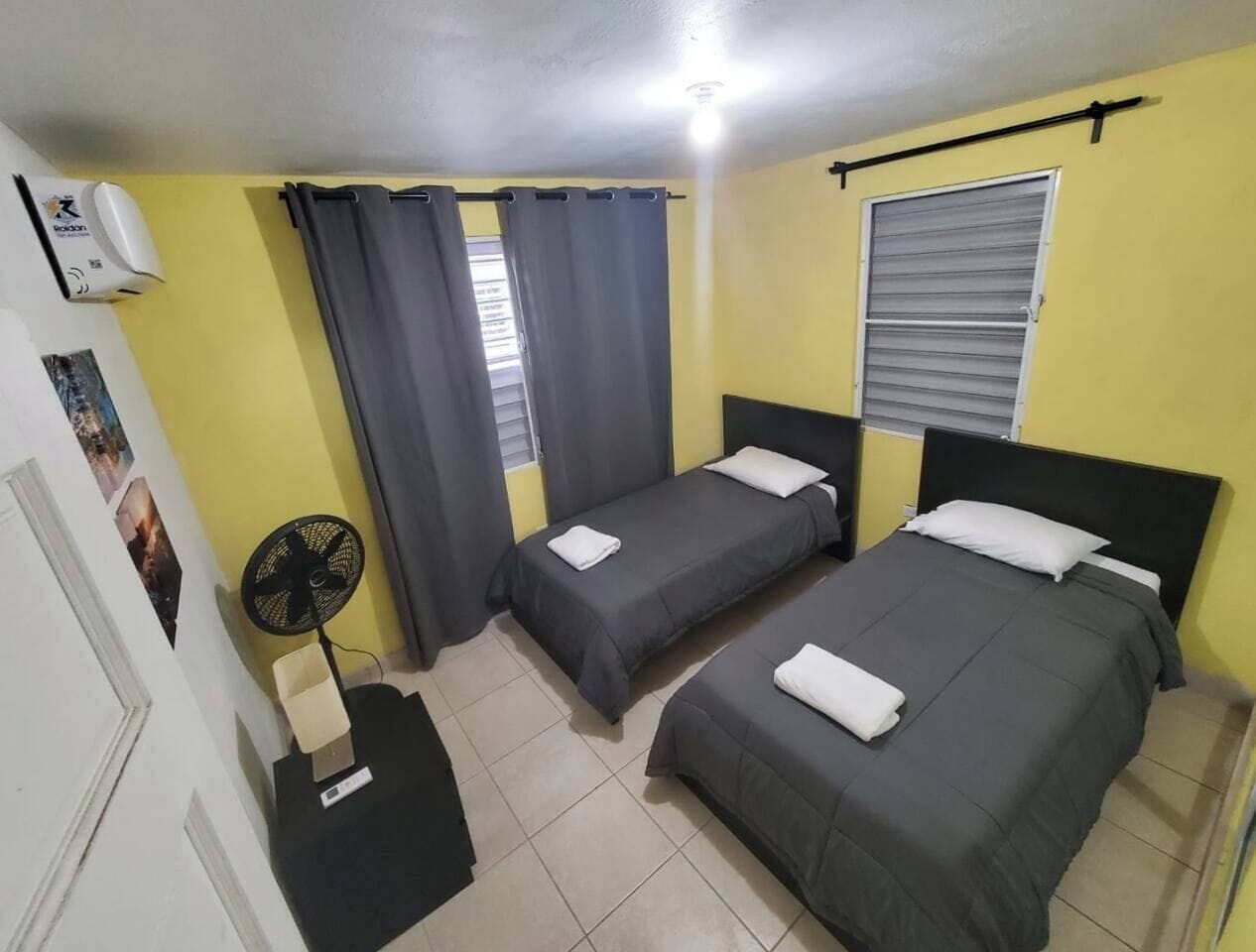 Une chambre avec deux lits, un ventilateur, une fenêtre avec des rideaux et une porte.
