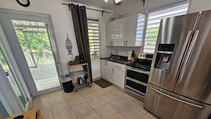 A kitchen with a stainless steel refrigerator, a sink, and a stove. There is a window with blinds, a door, and a small shelf with kitchen items.