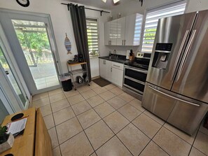 A kitchen with a stainless steel refrigerator, a sink, and a stove. There is a window with blinds, a door, and a small shelf with kitchen items. - Above the Plaza 7·Downtown Rincon·AC Rooms for 6 (Rincon)