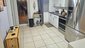 A compact kitchen with a stainless steel refrigerator, white cabinets, a small dining table, and a trash bin.