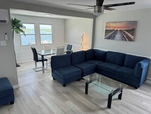 Ein modernes Wohnzimmer mit einem Ecksofa, einem Glastisch und Blick auf das Wasser durch große Fenster.