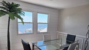 Een moderne kamer met een glazen tafel en stoelen, een hoge plant en uitzicht op zee door grote ramen.