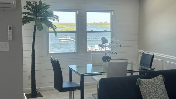 Een kamer met een glazen tafel, stoelen, een laptop, een bank, een palmboom en uitzicht op het water.