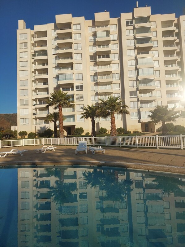 Un immeuble à plusieurs étages doté de balcons, d’une piscine avec des chaises longues et de palmiers.