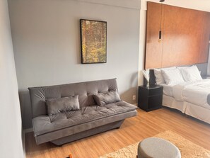 Une chambre moderne avec un canapé gris, un lit avec une literie blanche, une armoire en bois et un tableau encadré au mur.