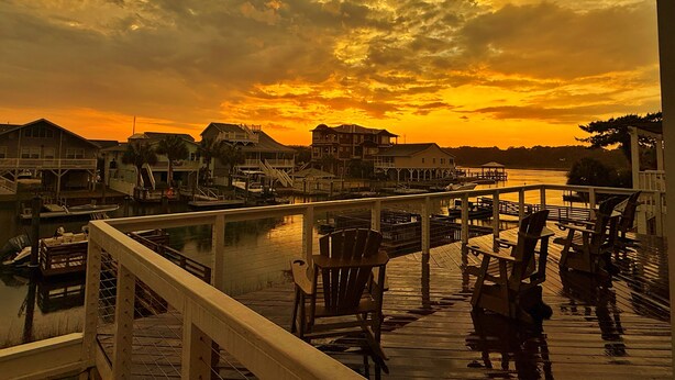 A waterfront view with a wooden deck, chairs, and a railing overlooking a marina and buildings. - Johnson's Coastal Cottage (Ocean Isle Beach)