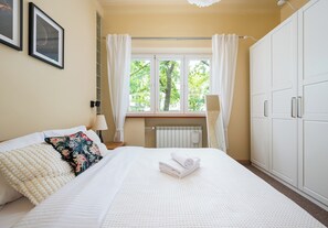 Ein Schlafzimmer mit Bett, Fenster mit Vorhängen, Heizkörper und Kleiderschrank.