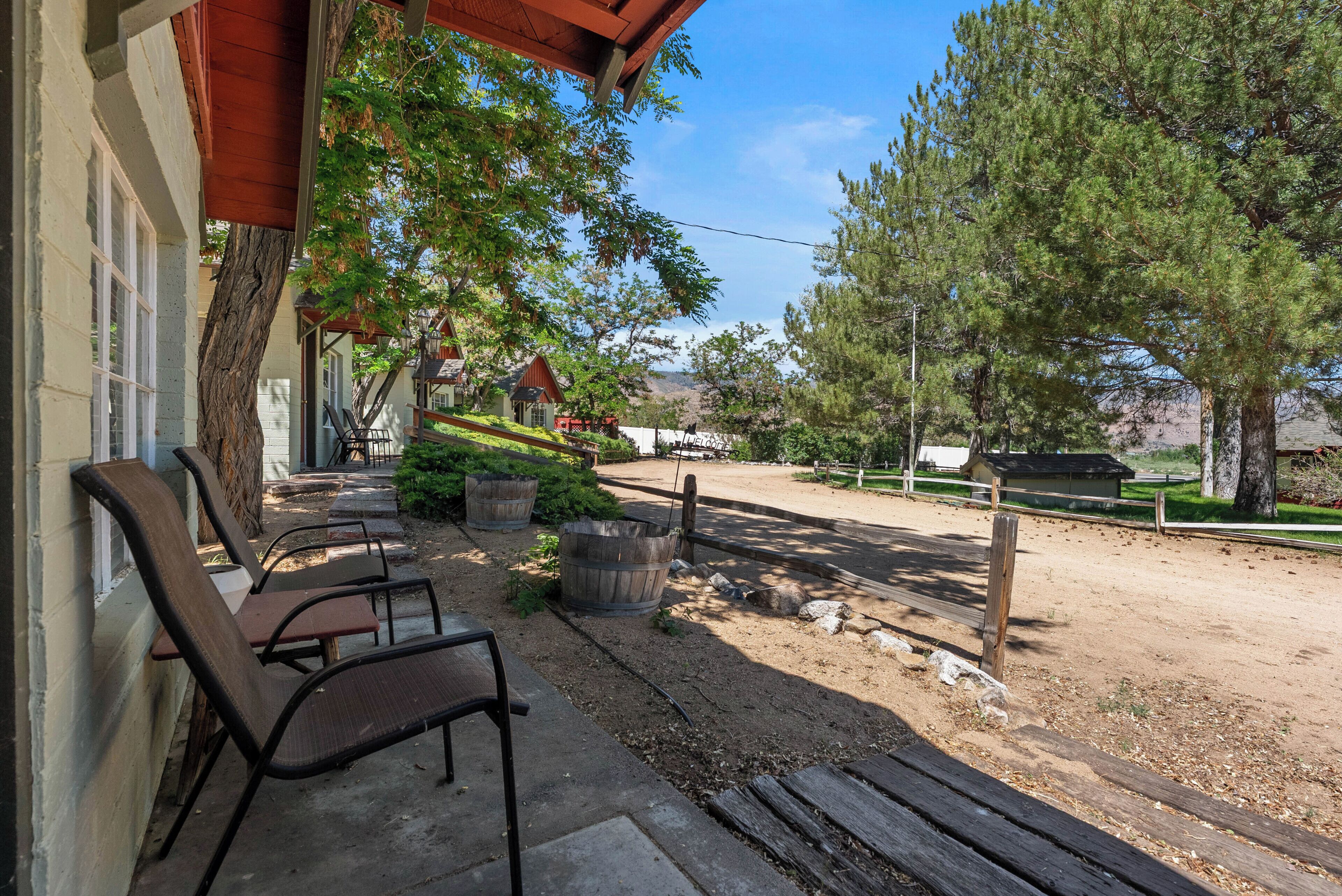 A building with a porch, outdoor seating, and a dirt path leading to a fenced area with trees.