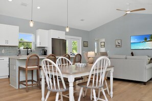 Une cuisine moderne avec des meubles blancs, une table à manger avec des chaises, un ventilateur de plafond et un grand téléviseur diffusant une scène de plage.