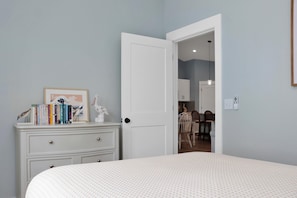 A bedroom with a bed, a dresser with books, a door leading to a dining area, and a wall-mounted light switch. - Pelican Point (Port St. Joe)