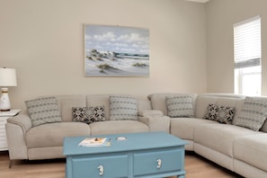 Ein Wohnzimmer mit einem beigefarbenen Sofa, gemusterten Kissen, einem blauen Couchtisch und einem Bild von einer Strandlandschaft.