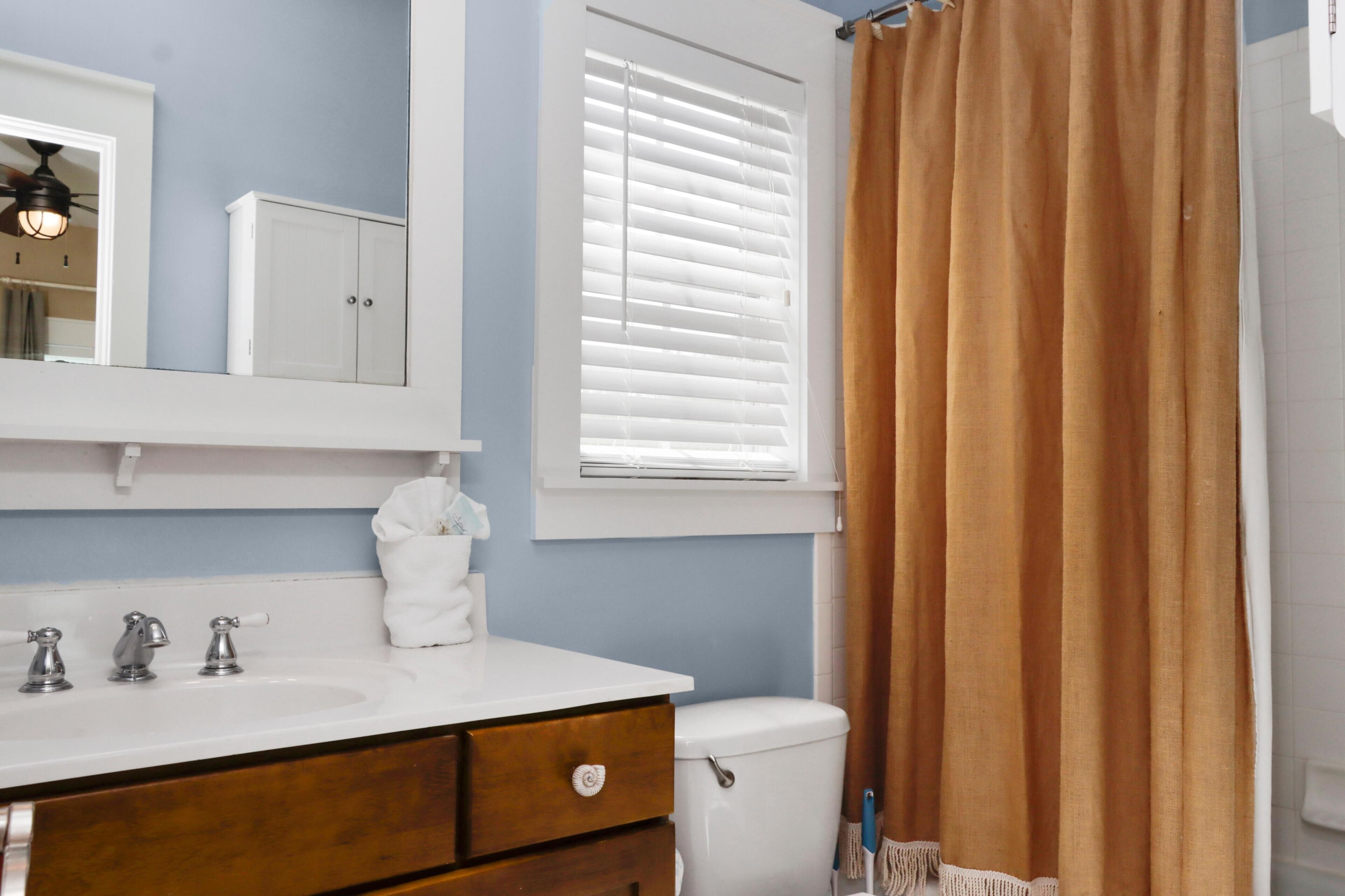 Une salle de bain avec un lavabo blanc, un miroir, un meuble de rangement, une fenêtre avec des stores et un rideau de douche.