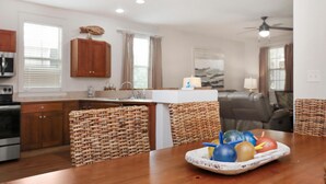 A modern kitchen with wooden cabinets, a dining table with wicker chairs, and a fruit bowl on the table. - Barefoot Cottage B28 (Port St. Joe)