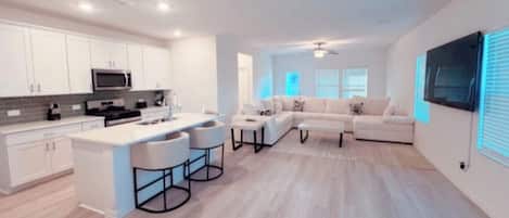 A modern kitchen with white cabinets, a central island with bar stools, a dining area with a white table and chairs, and a living area with a sofa and a TV mounted on the wall.