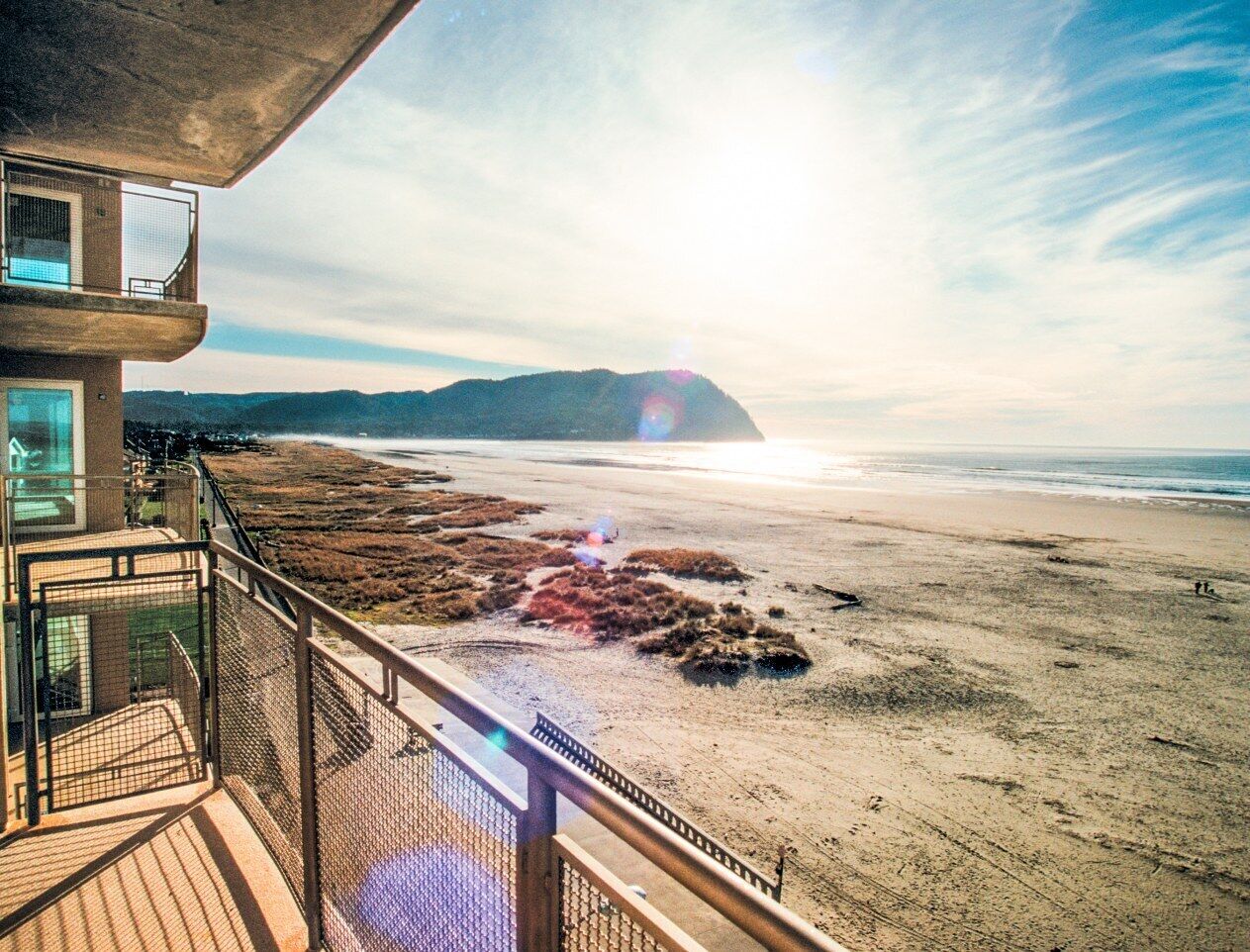 Une vue sur la plage depuis un balcon, avec un ciel dégagé, une plage de sable et des montagnes à l’horizon.