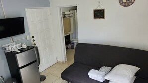 Ein Zimmer mit einer Couch, einem kleinen Kühlschrank, einem Fernseher, einer Tür zu einem Badezimmer und einer Wanduhr.