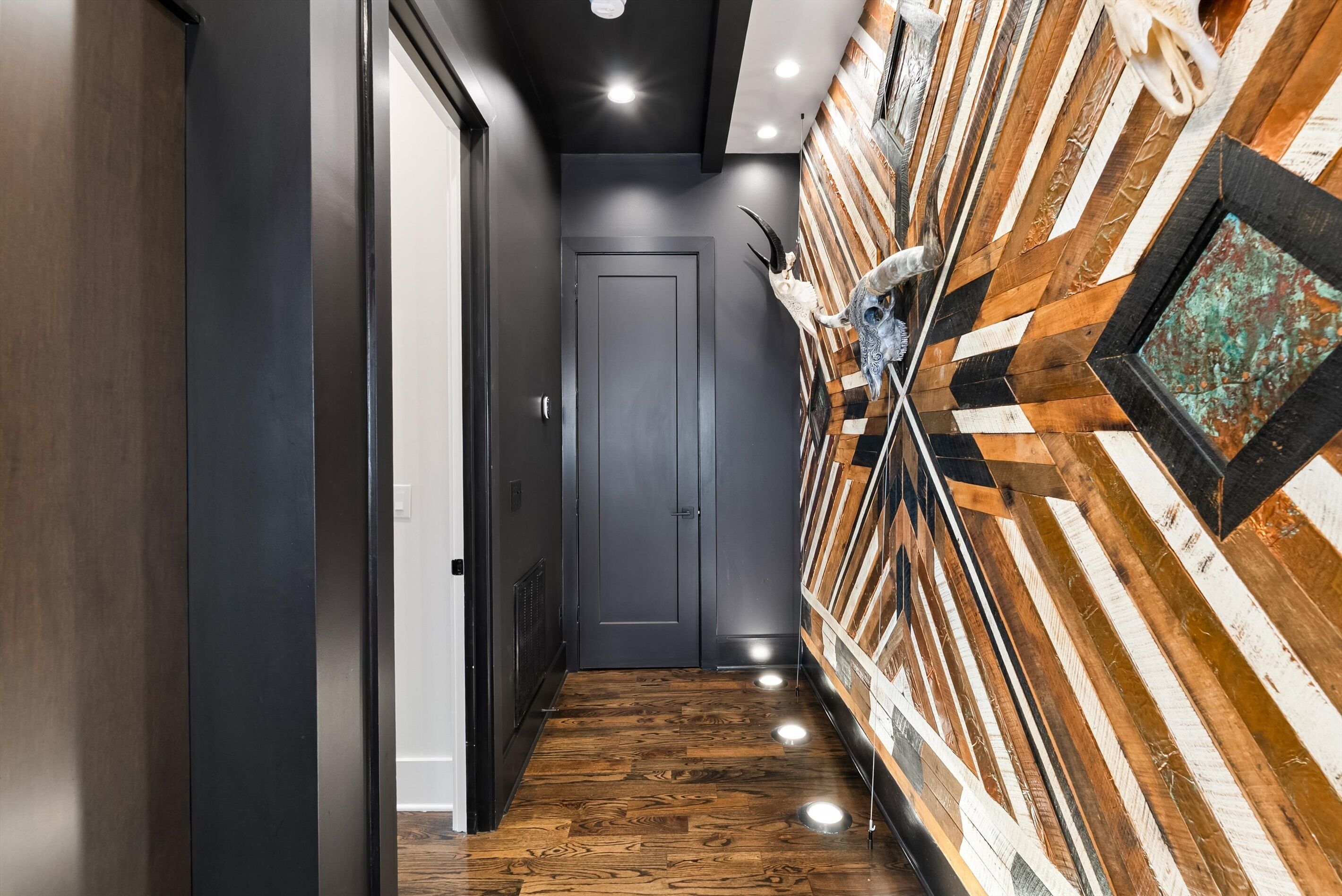Un corridoio con pavimento in legno, una porta grigia e un muro decorato con motivi geometrici e un teschio.