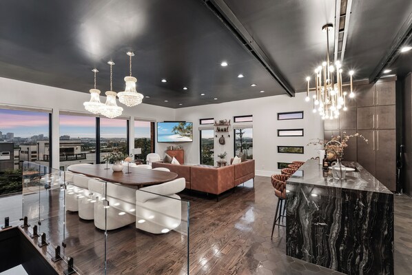 A modern living room with a glass dining area, a brown leather sofa, and a large TV.