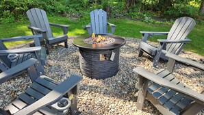 A fire pit surrounded by Adirondack chairs on a gravel surface.