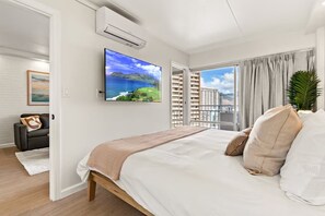 Ein modernes Hotelzimmer mit einem großen Bett, einem an der Wand befestigten Fernseher, einer Klimaanlage, Blick auf hohe Gebäude und einer Zimmerpflanze.