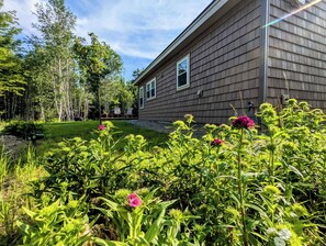 A house with a deck, surrounded by lush greenery and flowering plants. - New Garden Retreat w/ Fire Pit, 5 Min to Town (Ellsworth)