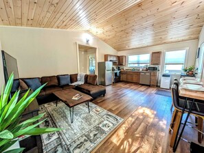 A spacious living area with a sofa, coffee table, and a rug. A kitchen with wooden cabinets and a dining area with a table and chairs is visible. - New Garden Retreat w/ Fire Pit, 5 Min to Town (Ellsworth)
