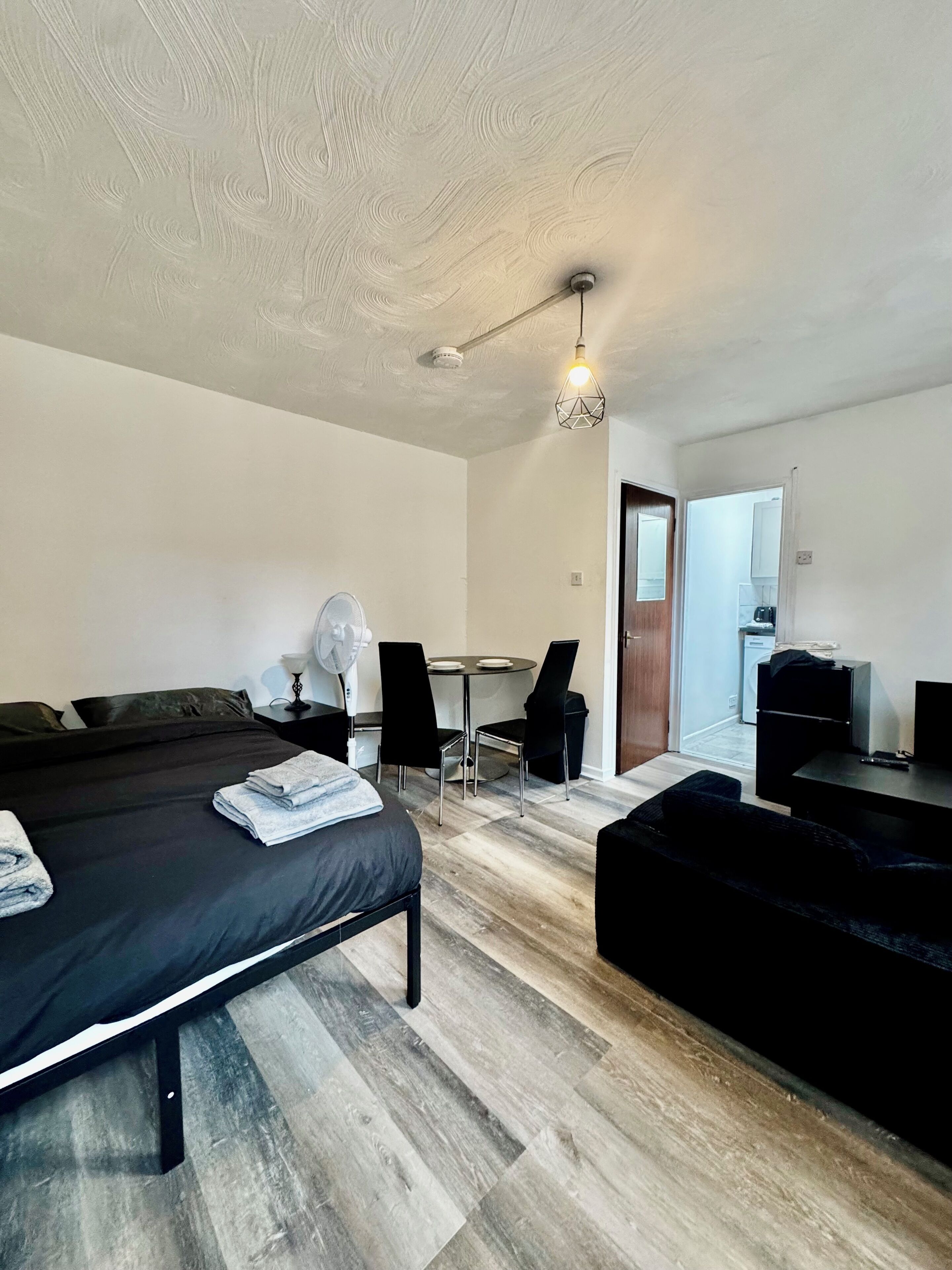 A modern hotel room with a bed, a small table with chairs, a kitchenette, and a door leading to another room.