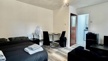 A modern hotel room with a bed, a small table with chairs, a kitchenette, and a door leading to another room.